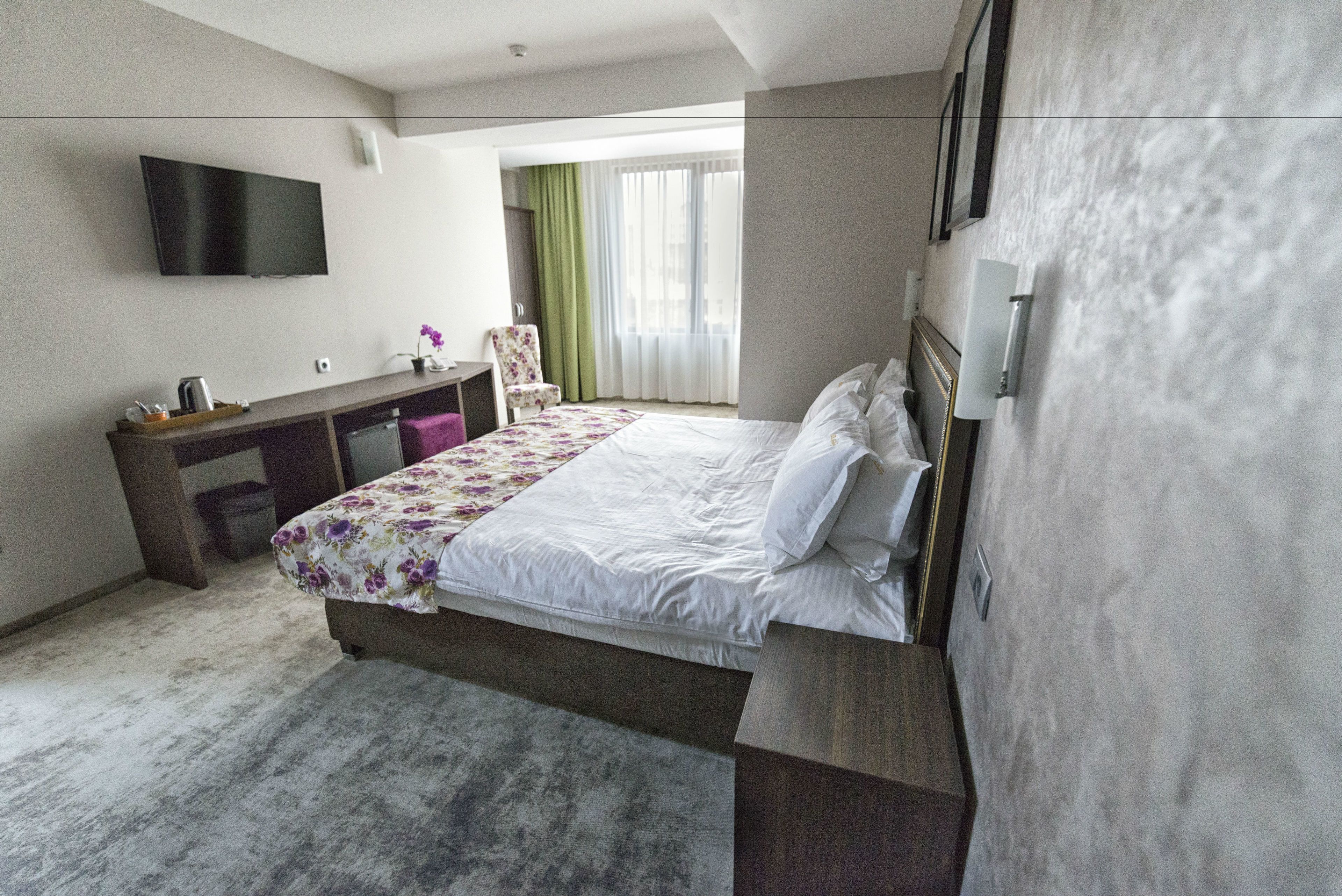 A modern hotel room with a bed, a desk, a chair, a flat-screen TV, and a large window with curtains.
