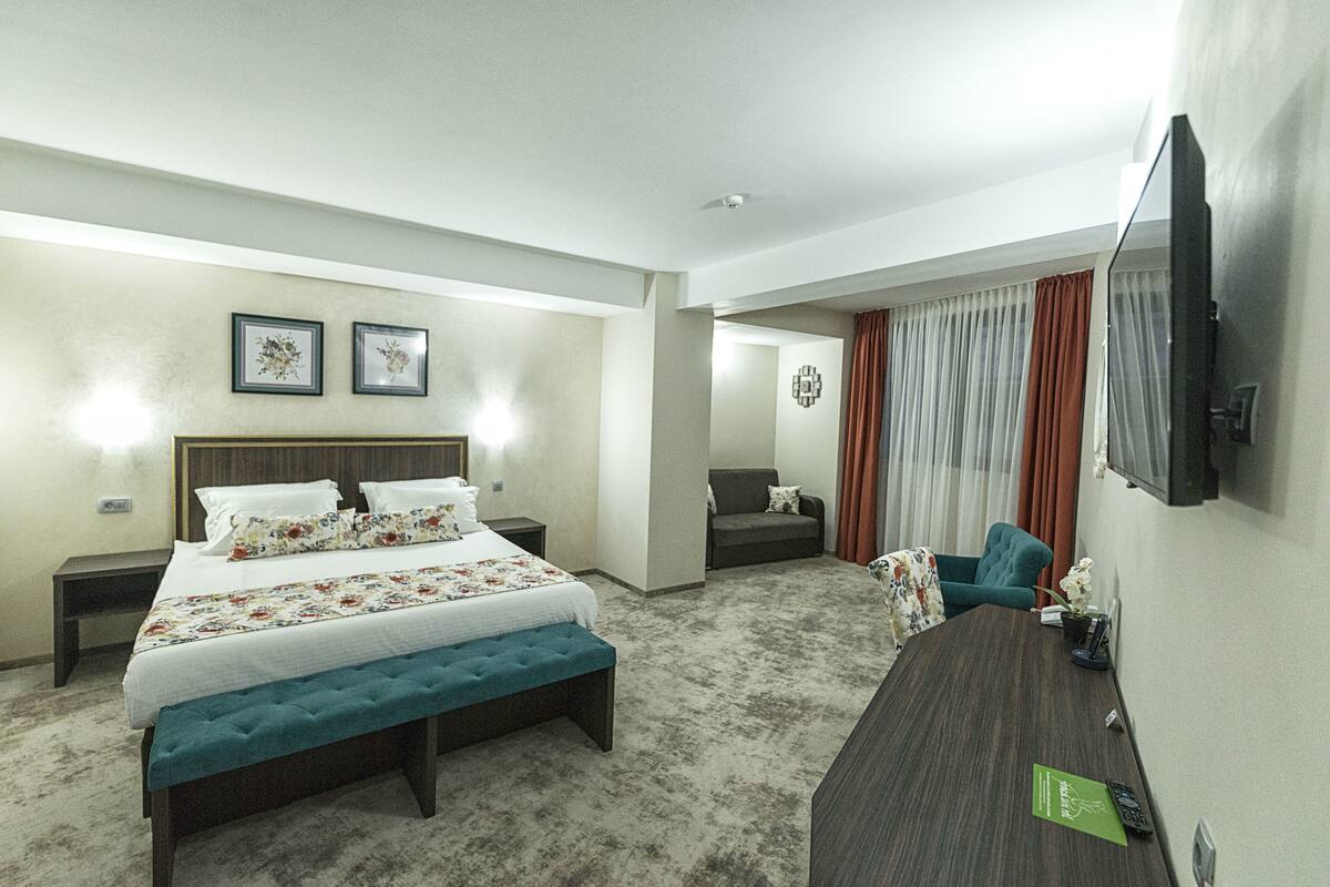 Habitació d'hotel amb un llit, una banca, escriptori, cadira, televisió i finestra amb cortines.