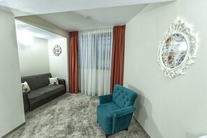 Una sala de estar con un sofá gris, un sillón azul aguamarina, un espejo estampado y una ventana con cortinas translúcidas.