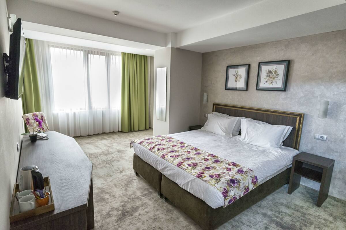 Una habitació d'hotel amb un llit gran, una taula de nit, una cadira, un escriptori i una finestra amb cortines.