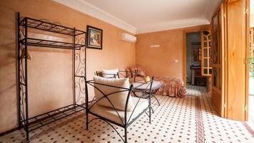 A room with a metal bed frame, a small table with a cushion, patterned floor tiles, and a wooden door.