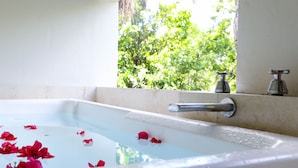 A bathtub with rose petals floating on the water, a window with a view of greenery, and two faucets on the wall. - Turquesa Penthouse 2 BDRM sleeps 5 (Tulum)