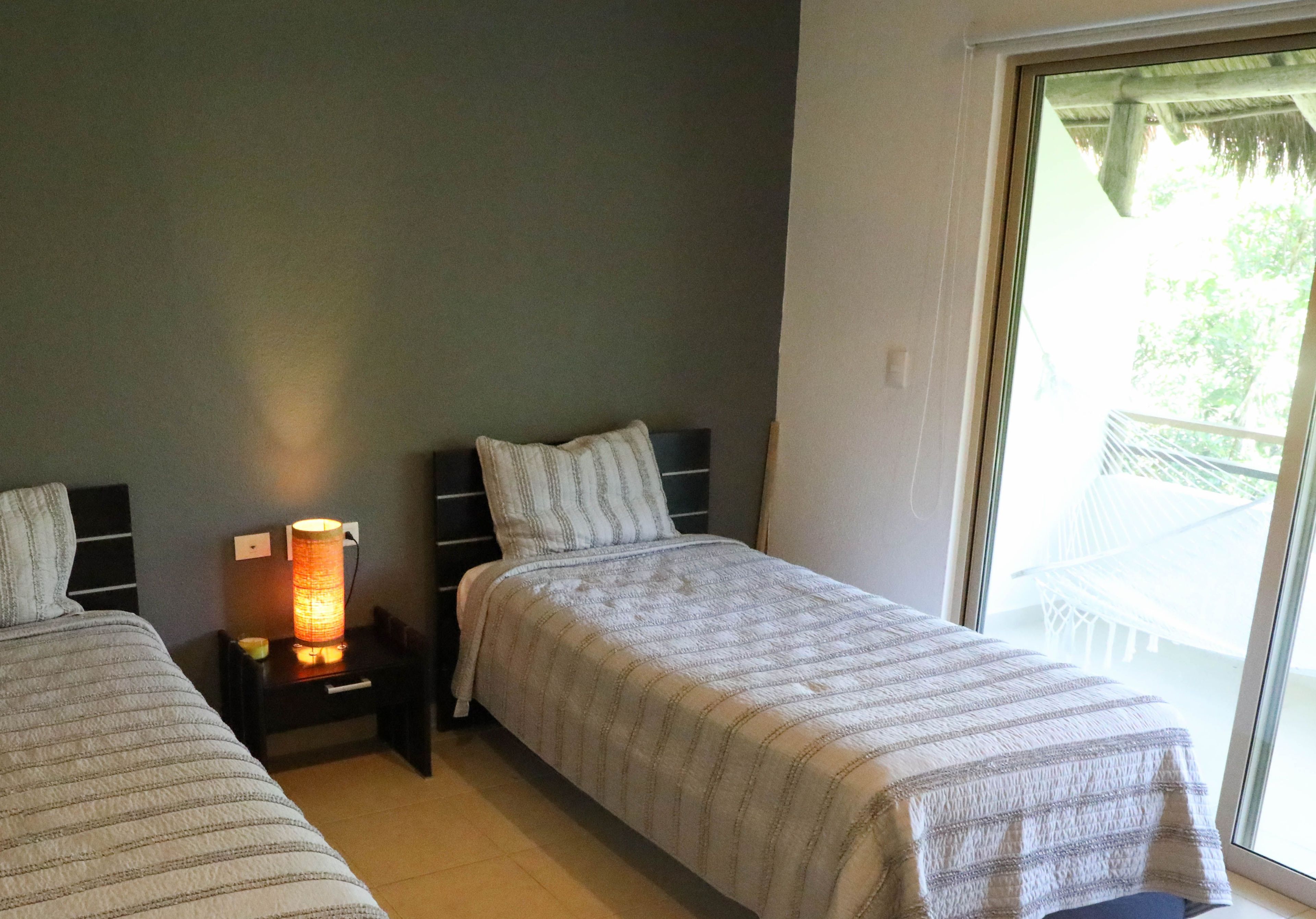 A bedroom with two beds, a nightstand with a lamp, a window with a view of an outdoor area.