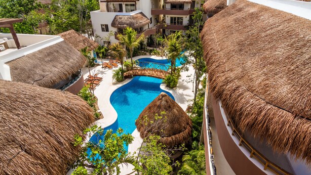 Resort com edifícios de telhado de palha, piscina central e vegetação exuberante.