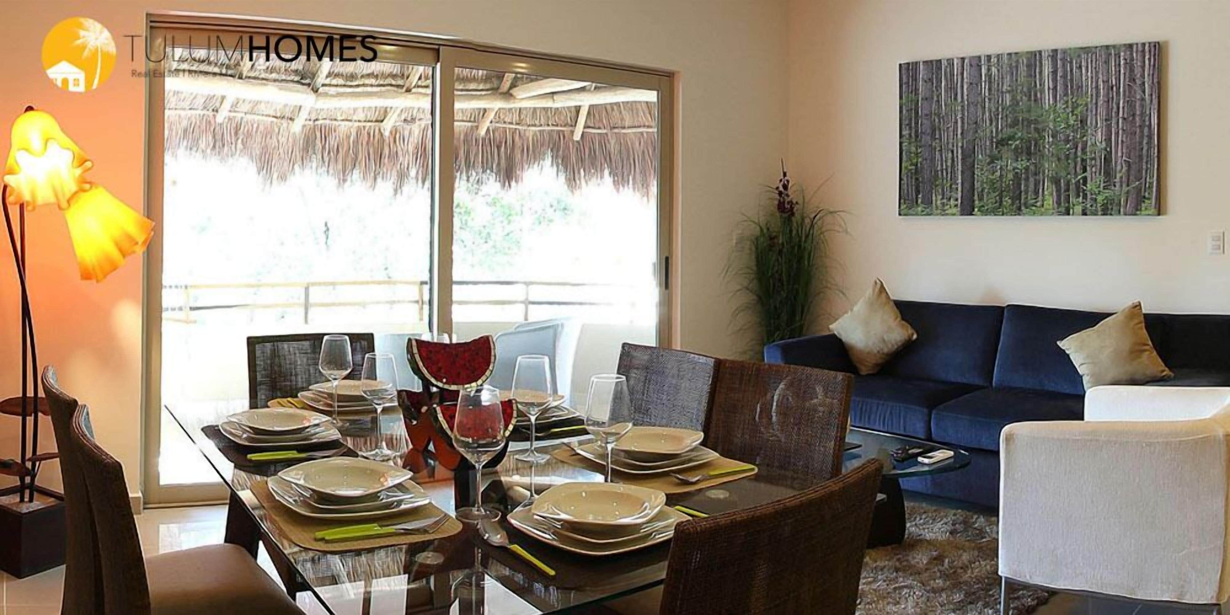 A dining area with a glass table set for six, a blue sofa, and a painting of a forest on the wall.