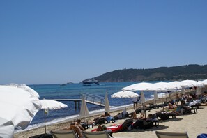 Una spiaggia con lettini e ombrelloni, un molo e barche sullo sfondo.