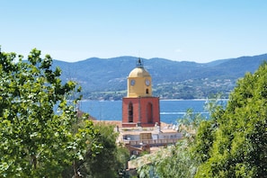 Un edificio costero con una torre de reloj, rodeado de vegetación y con vista a una masa de agua con montañas al fondo.