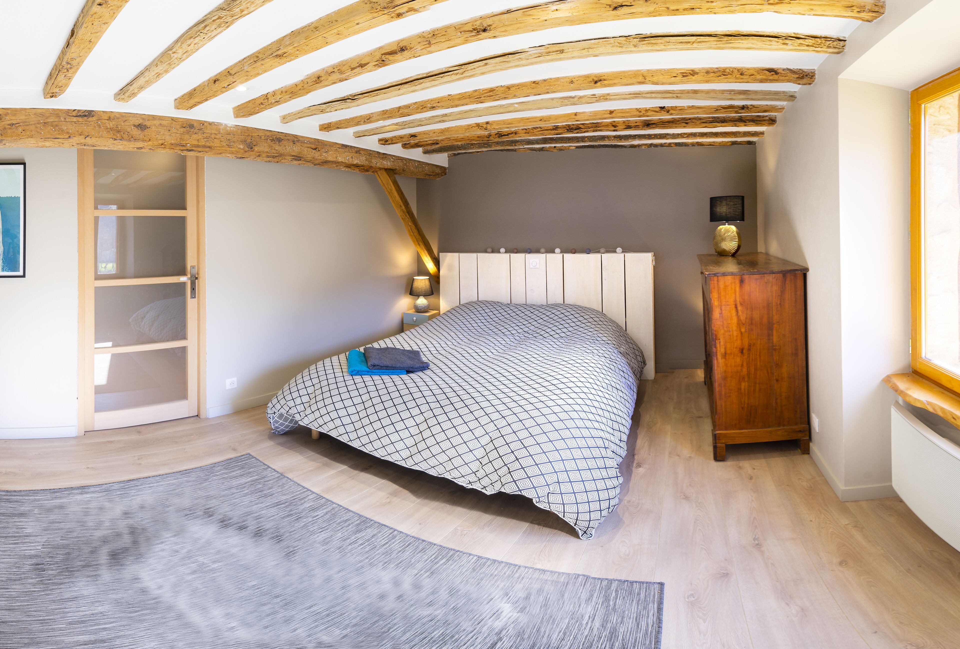 Un dormitorio con techo de madera, una cama con colcha estampada, una mesita de noche con lámpara y una mesita auxiliar de madera.