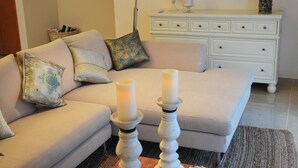 Een woonkamer met een beige bank, een houten salontafel met kaarsen en decoratieve objecten, een witte dressoir en een spiegel met een sierlijk frame.
