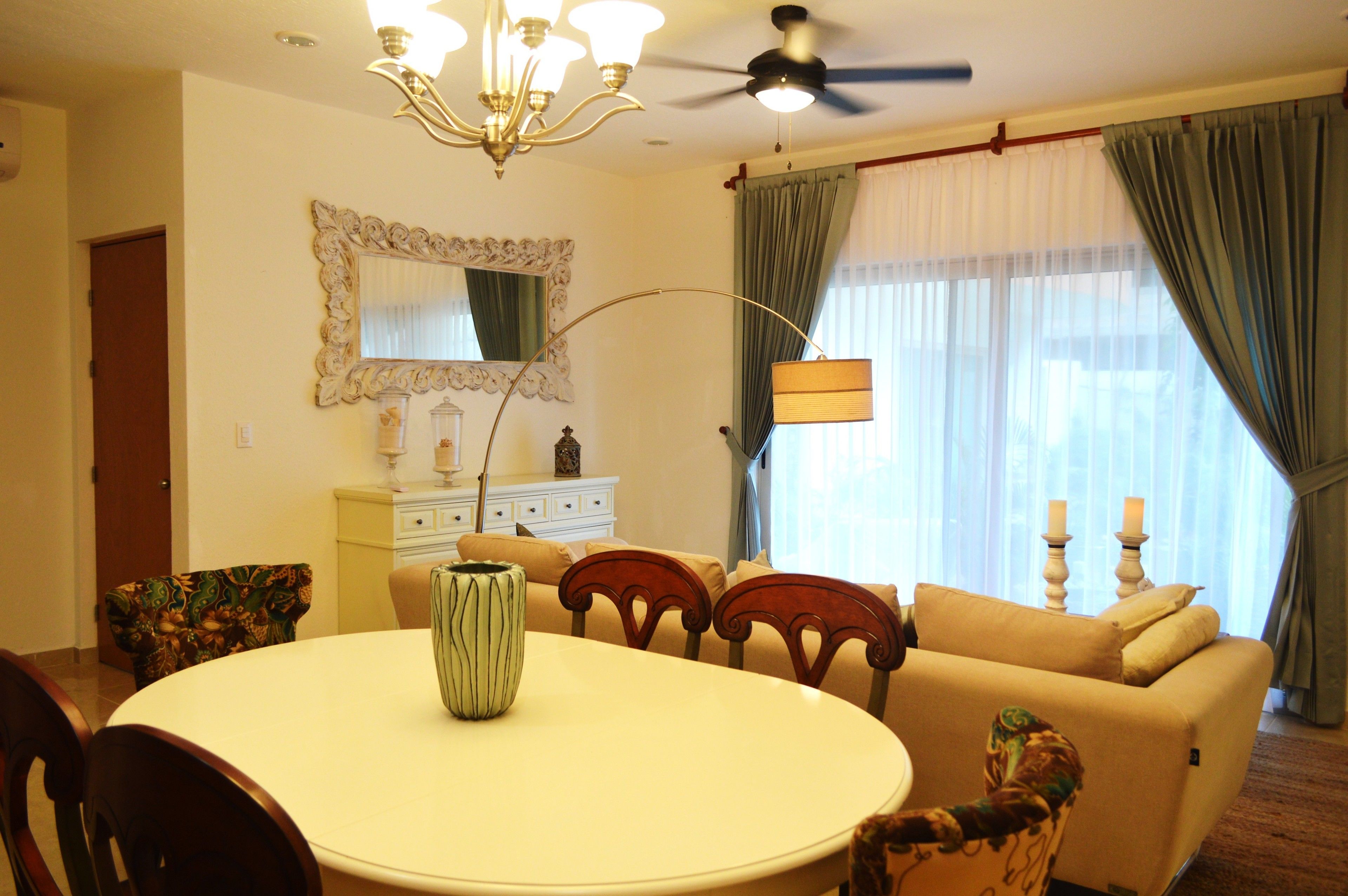 A dining area with a round table, chairs, a chandelier, a ceiling fan, and a window with curtains.
