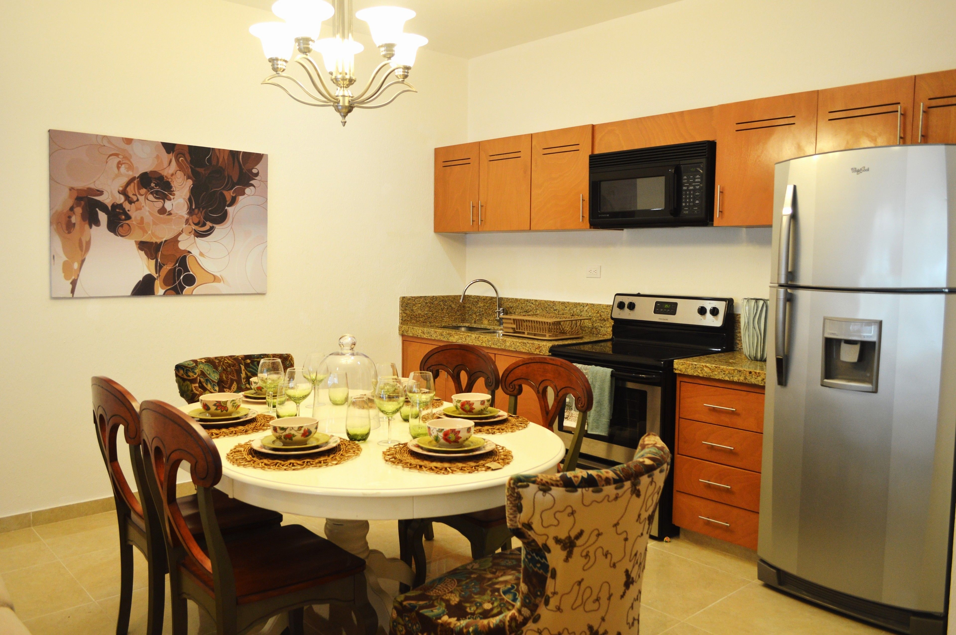 A dining area with a round table set for four, surrounded by wooden chairs. A chandelier hangs above the table, and a painting is on the wall.