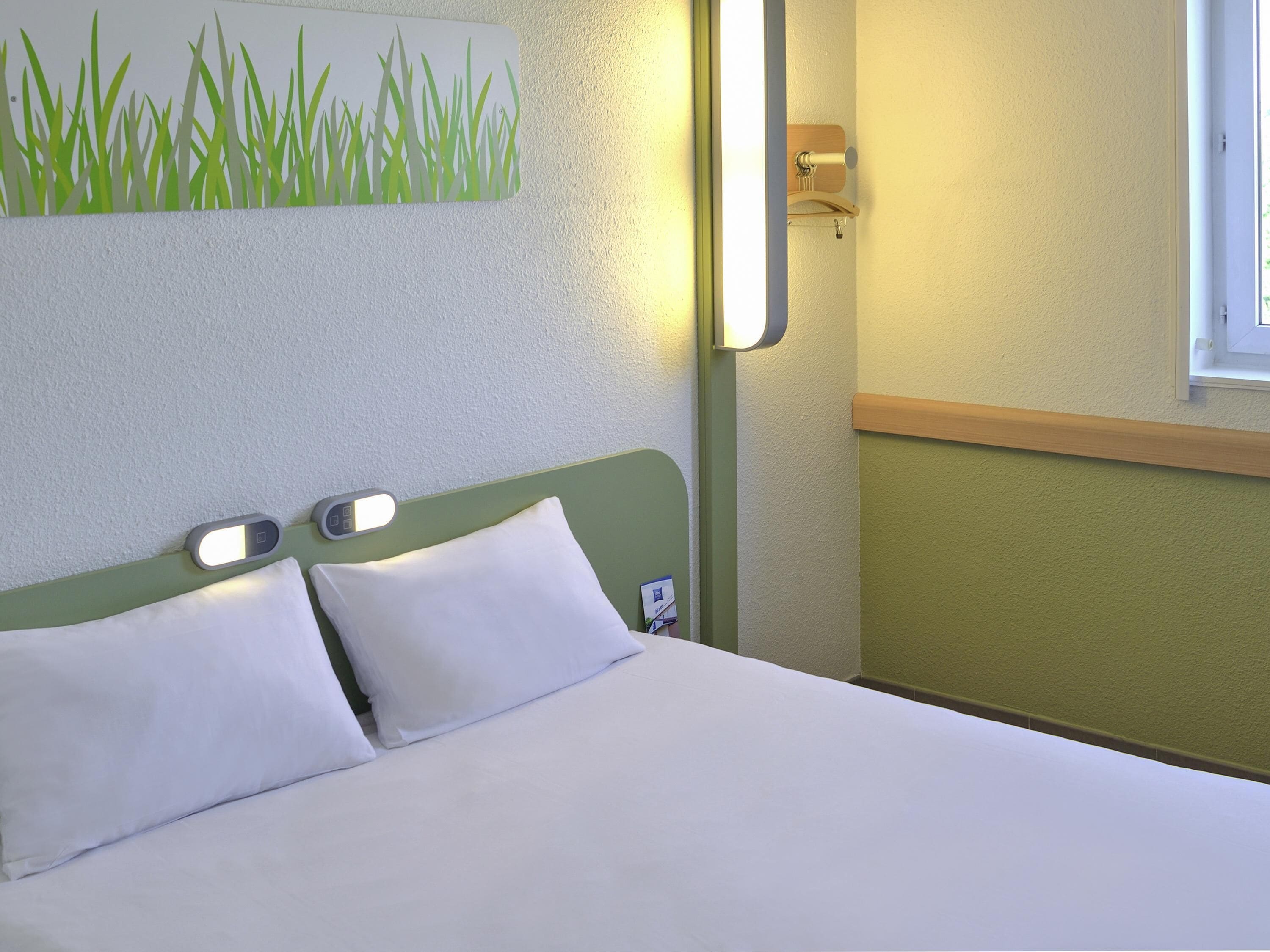 A neatly made bed with white linens, a green headboard, and a wall-mounted light fixture.