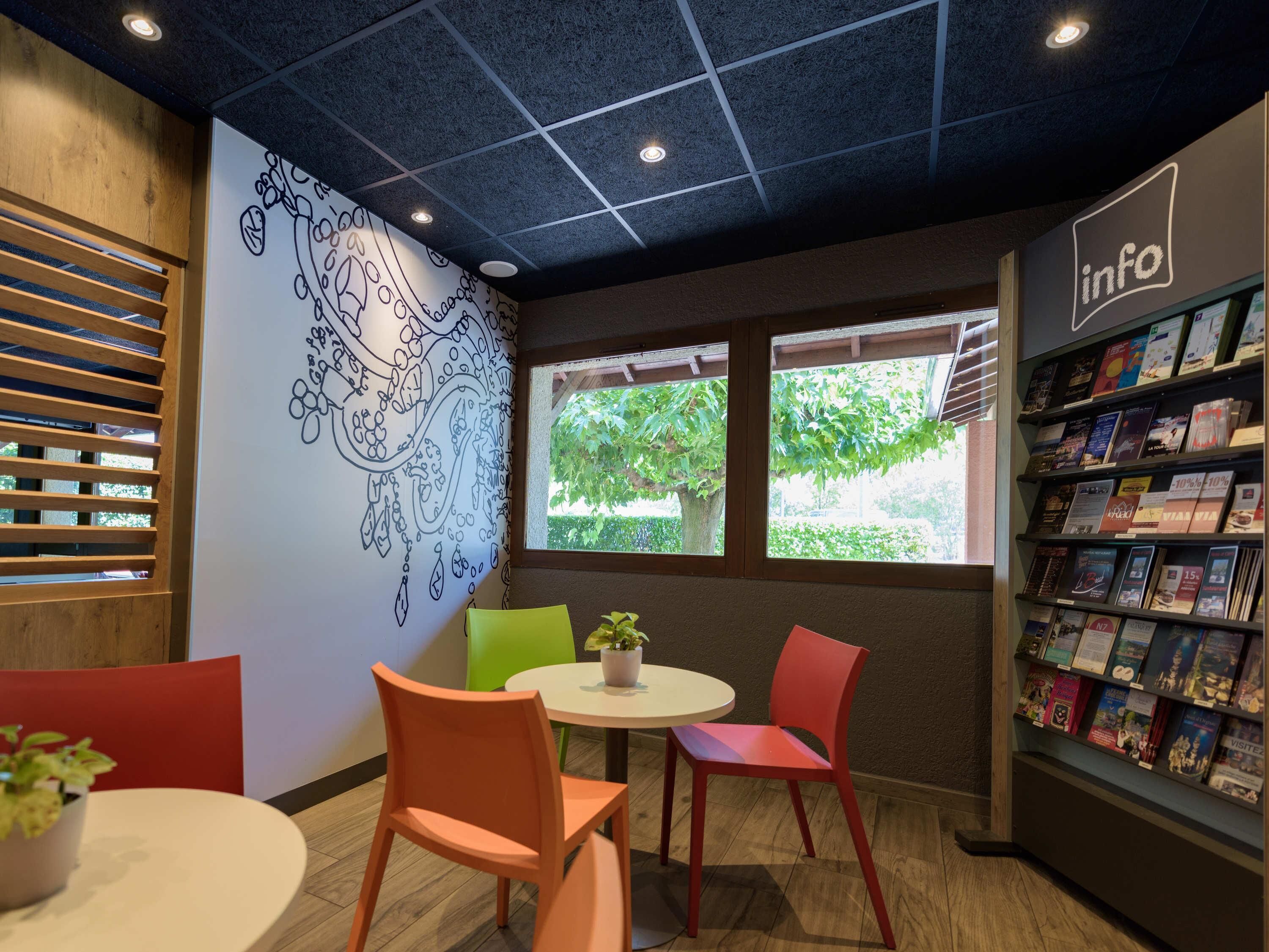 A cozy reading area with a mural, a bookshelf, and two windows with greenery outside.