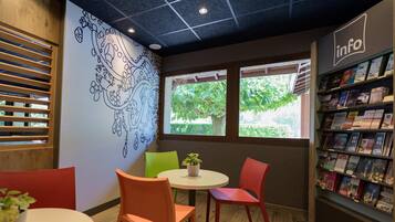 A cozy reading area with a mural, a bookshelf, and two windows with greenery outside.