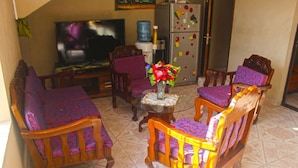 A living room with a TV, wooden chairs with patterned cushions, a small table with a vase of flowers, and a refrigerator with magnets. - Marcie Guesthouse hôtel and restaurant (Tabarre)