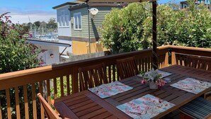 Eine Holzterrasse mit einem Tisch für zwei Personen, einem großen Sonnenschirm und Blick auf einen Yachthafen mit Booten und einem Haus im Hintergrund.
