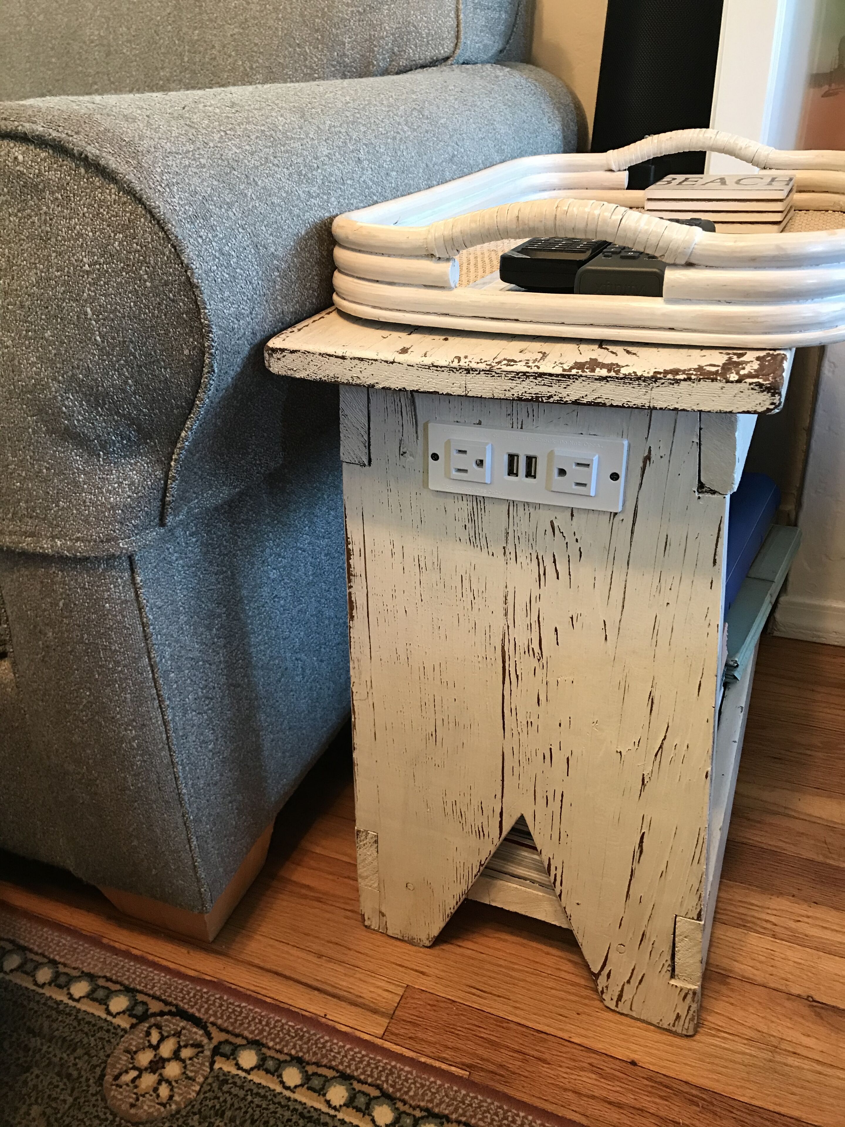Una mesa auxiliar de madera rústica con un enchufe, ubicada al lado de un sofá gris.