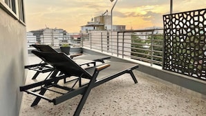 A rooftop terrace with two lounge chairs and a table, a decorative metal fence, and a view of the cityscape during sunset.