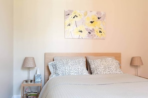 A bedroom with a bed, two bedside tables, a lamp on each table, a wall-mounted painting, and a bookshelf with books.