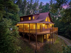 A two-story wooden cabin with a balcony, surrounded by dense forest.