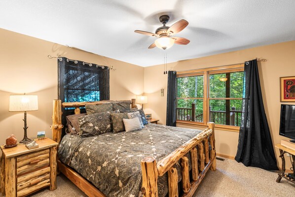 A bedroom with a wooden bed frame, a ceiling fan, a window with curtains, and a nightstand with a lamp.