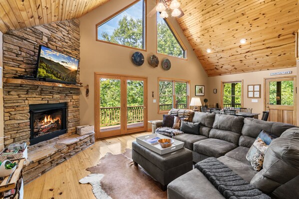 A spacious living room with a stone fireplace, leather couches, and large windows offering views of the outdoors.