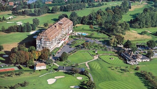 Eine Vogelperspektive auf einen Golfplatz mit einem großen Hotelgebäude, mehreren Grünflächen und einem Schwimmbad.