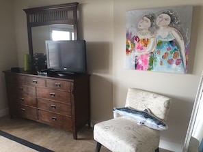 Een kamer met een houten dressoir, een televisie erboven, een stoel met een kussen en een schilderij aan de muur.