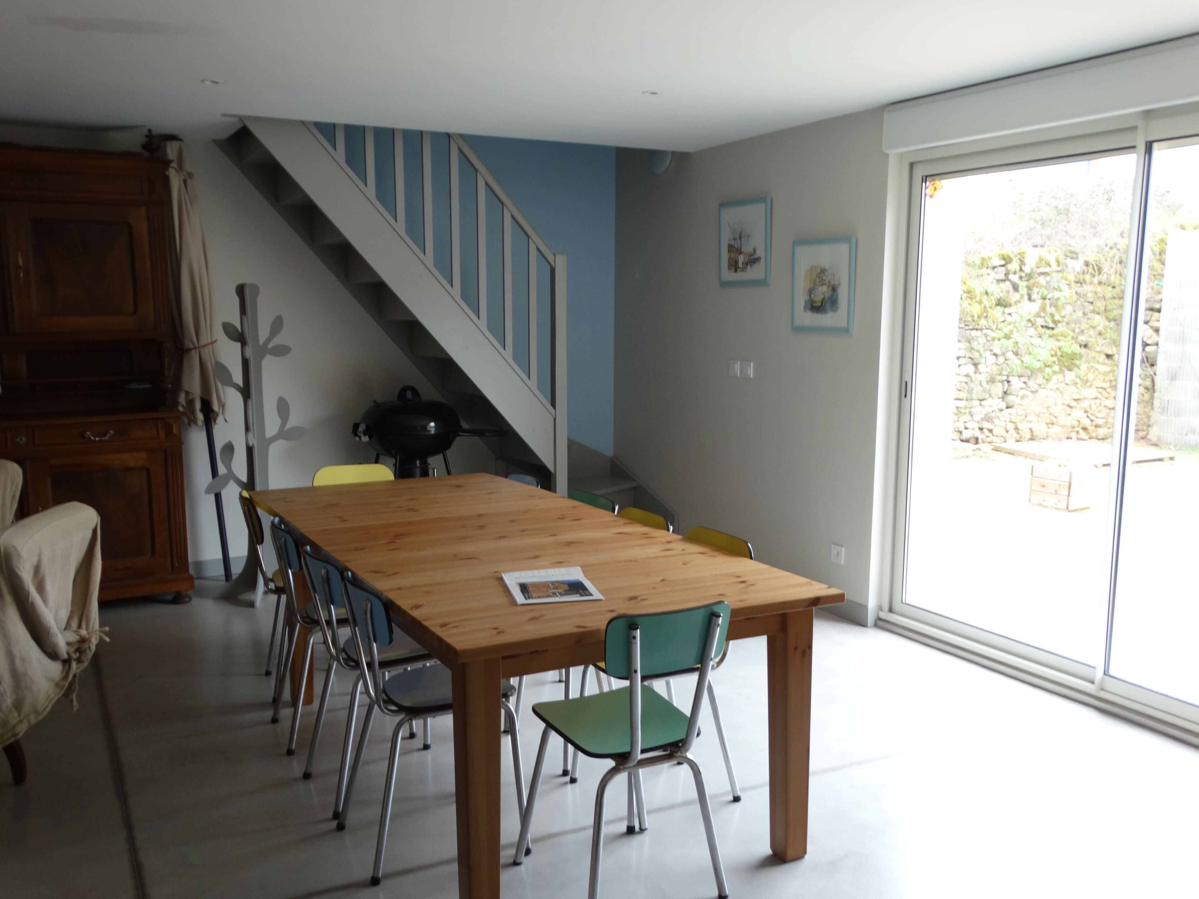 Un coin repas avec une table et des chaises en bois, un escalier et une vue dégagée sur l’extérieur grâce à une grande fenêtre.