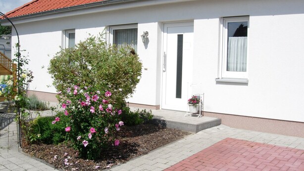 A white building with a red-tiled roof, a small garden with pink flowers, and a paved walkway.