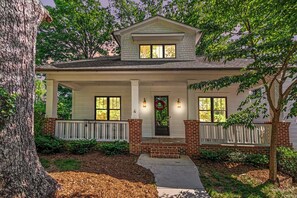 A two-story house with a front porch, brick foundation, and a large tree in the foreground.
