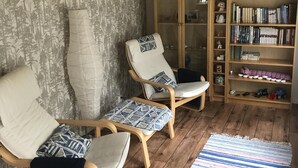 A room with three wooden chairs with cushions, a bookshelf filled with books, and a patterned rug on the floor. - Seglarvägen 18 - House in south-east Sweden - Oknö, Mönsterås (Oknö)