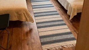 A small, cozy bedroom with two single beds, a wooden floor, a striped rug, a window with curtains, and a view of a house outside. - Seglarvägen 18 - House in south-east Sweden - Oknö, Mönsterås (Oknö)