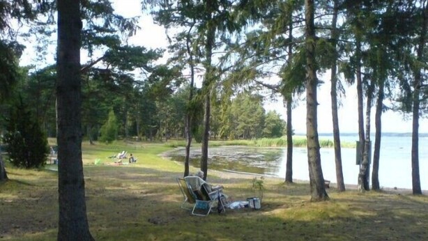 A campsite with a chair and camping gear, surrounded by trees and a lake. - Seglarvägen 18 - House in south-east Sweden - Oknö, Mönsterås (Oknö)