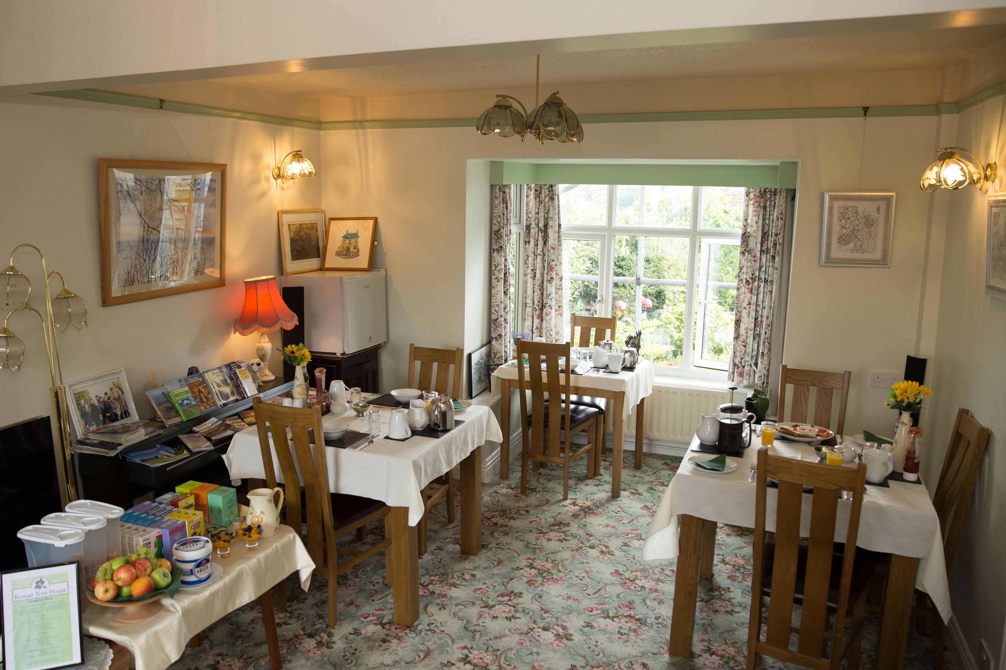A dining area with wooden tables and chairs, a window with floral curtains, and various dining items on the tables.