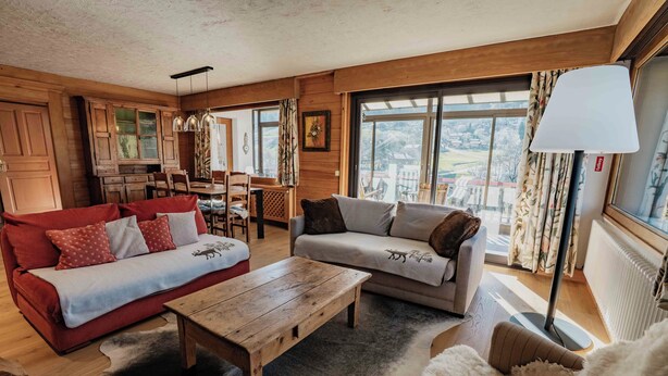 A cozy living room with a wooden ceiling, a dining area with a table and chairs, and a large window with a view.