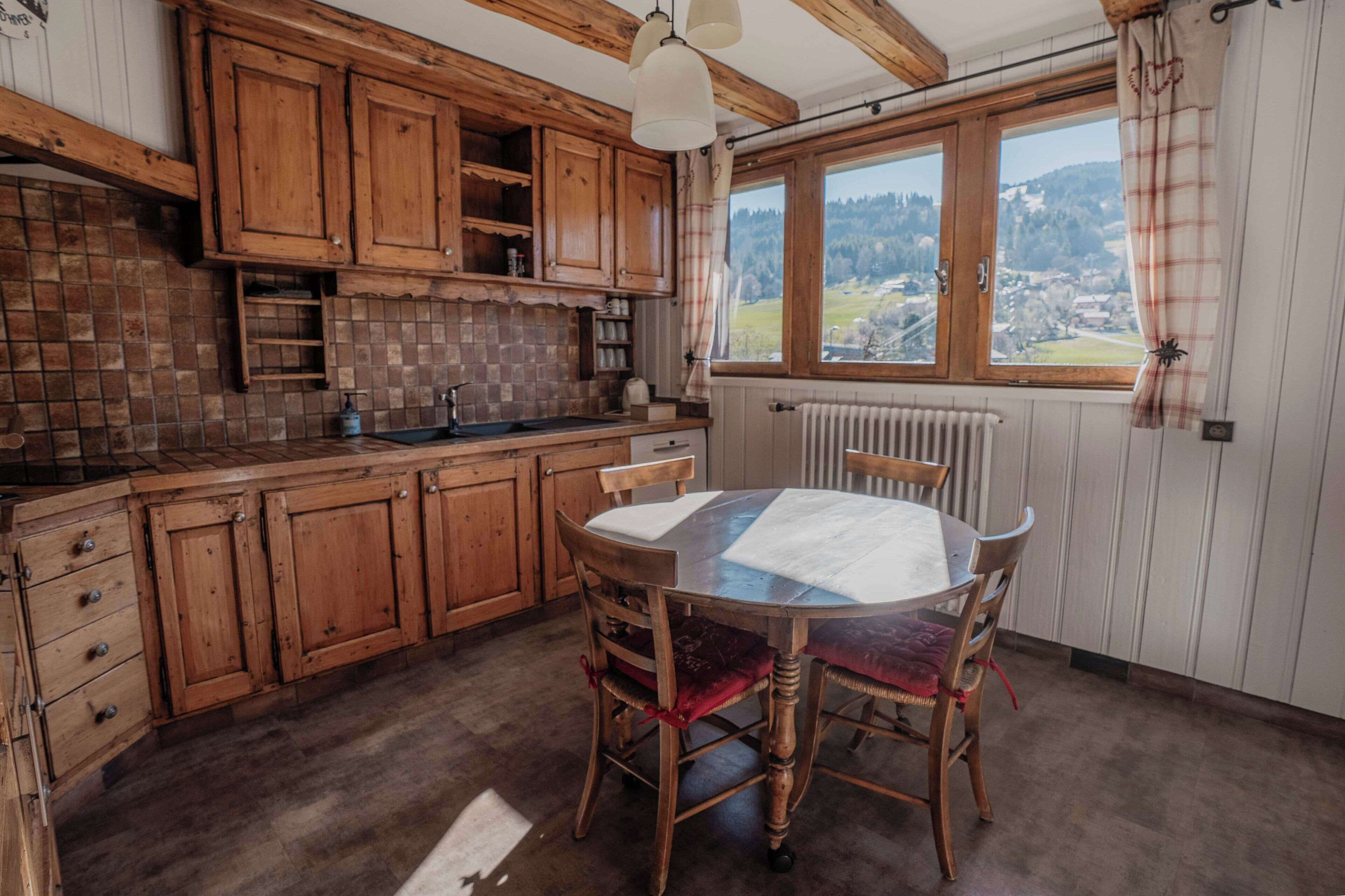 A kitchen with wooden cabinets, a round table with chairs, and a view through a window.