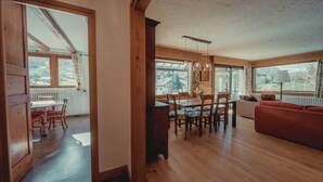 A spacious living area with wooden flooring, a dining table with chairs, a sofa, and large windows offering views of the outdoors.