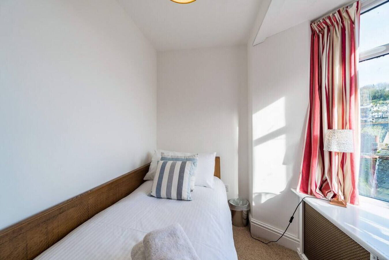 A single bed with a wooden headboard, white bedding, and striped pillows. A window with red and white curtains allows natural light into the room.