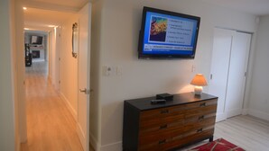 A hotel room with a television displaying an event schedule, a wooden dresser, and a red rug. - Marquise 204 Beachfront 2br, 2 Bath condo-Directly on Beach! (Indian Rocks Beach)