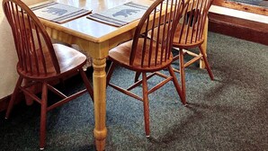 A dining area with a wooden table, four chairs, a decorative bear figurine, and a window with patterned curtains. - Wilderness Spirit Cabins 'Bear Cave' cabin-family friendly/sportsman paradise (Corvallis)