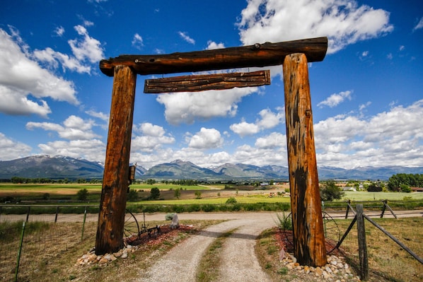 Un arco de madera rústico con un camino de grava que lo atraviesa, con montañas y un cielo despejado al fondo.