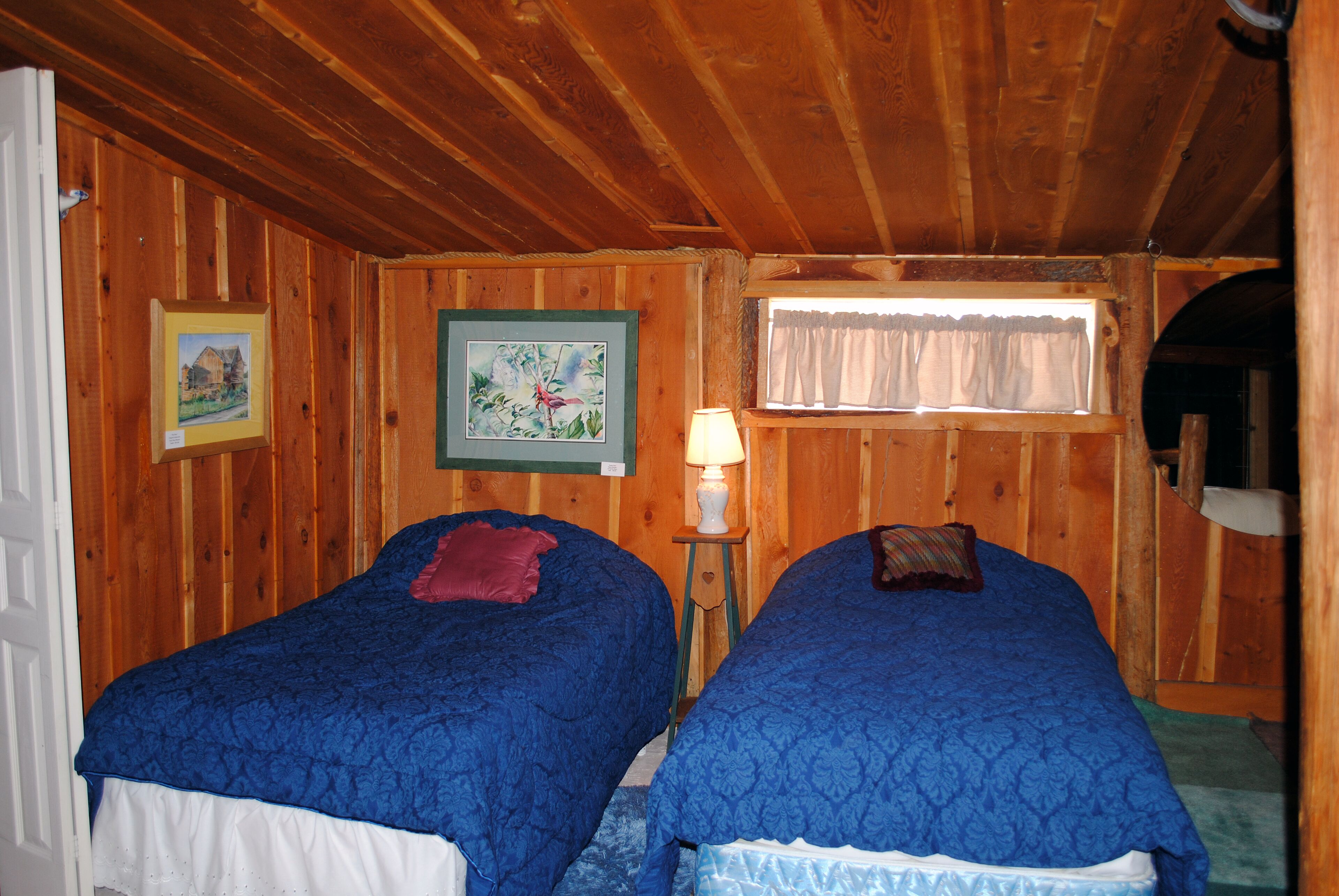 A room with wooden walls and two beds with blue bedding. There is a framed picture on the wall and a window with curtains.