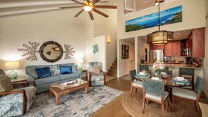 A spacious living room with a high ceiling fan, a dining area with a round table, and a kitchen with wooden cabinets. - SPECIAL SUMMER BREAK RATES! Stunning Ocean View 2BR/2BA condo across KAM3 Beach! (Kihei)