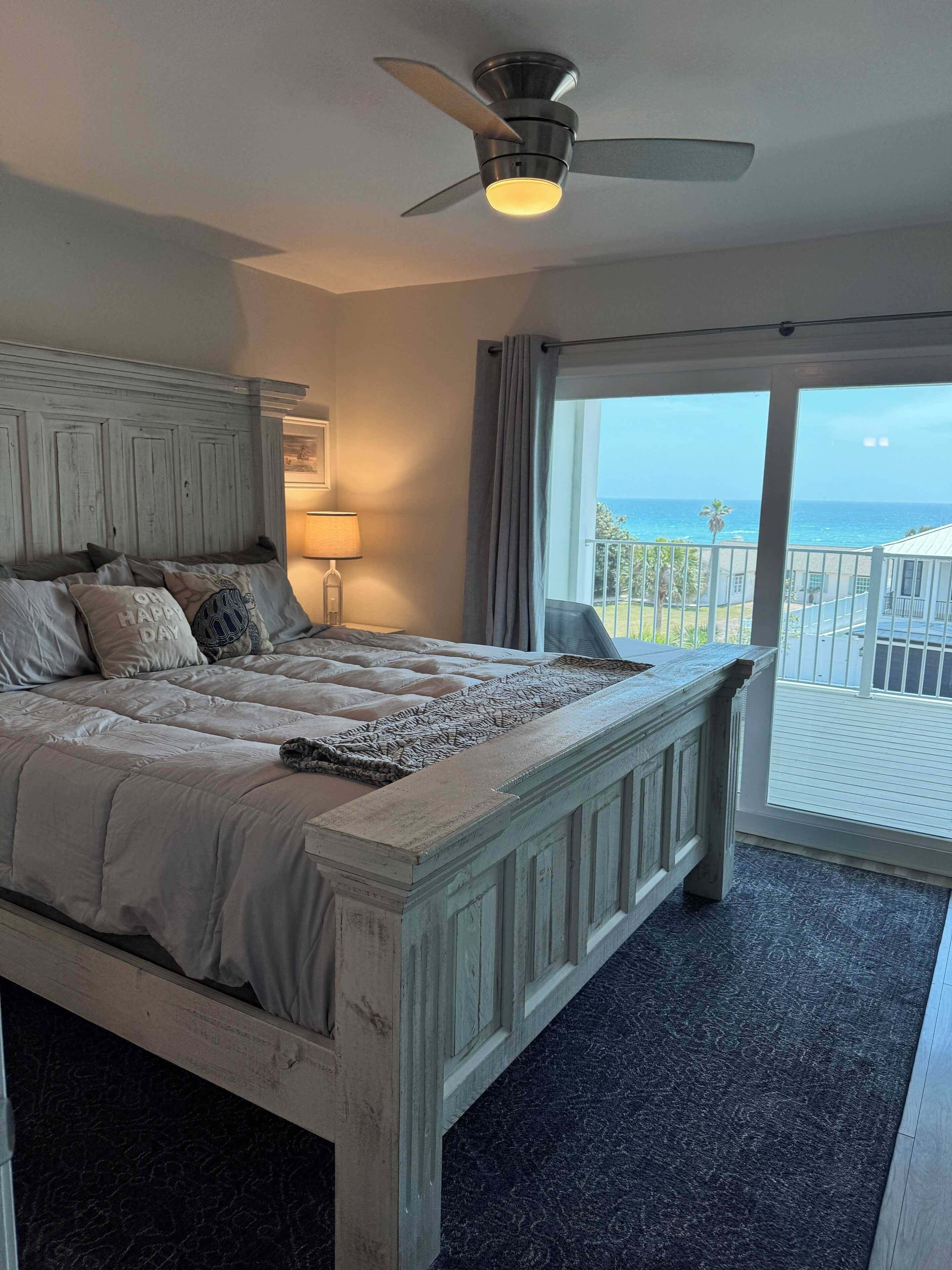 A bedroom with a large bed, a ceiling fan, a view of the ocean, and a balcony.