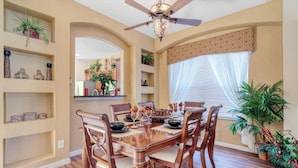 A dining area with a wooden table set for six, surrounded by chairs. There is a ceiling fan above and a window with sheer curtains to the right.