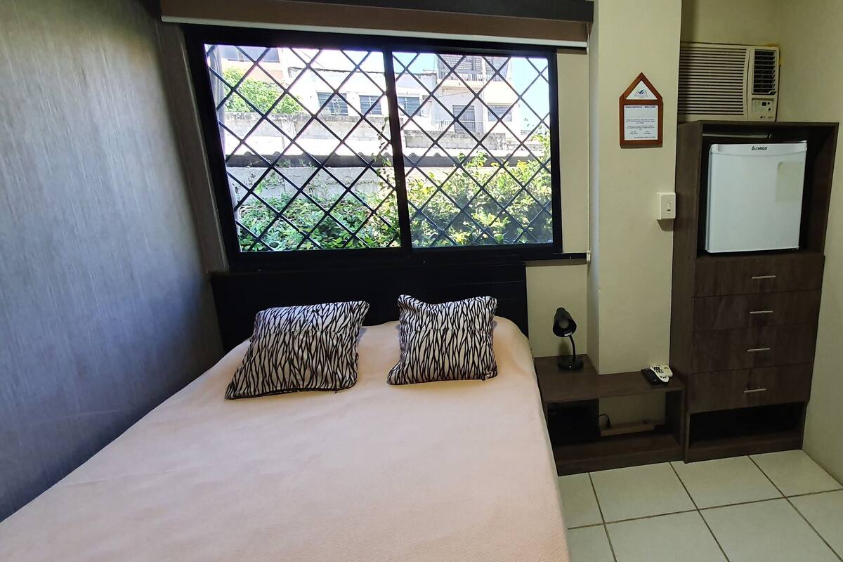 A bedroom with a bed, patterned pillows, a nightstand with a lamp, a wooden wardrobe, a window with a view of buildings and greenery, and a painting on the wall.