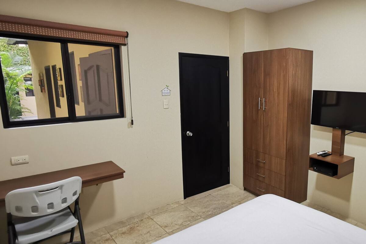 A bedroom with a bed, a wooden wardrobe, a desk with a chair, a television, and a bathroom with a sink and mirror.