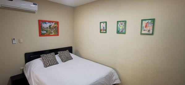 Habitación de hotel con cama, dos unidades de aire acondicionado fijadas a la pared y tres cuadros enmarcados.
