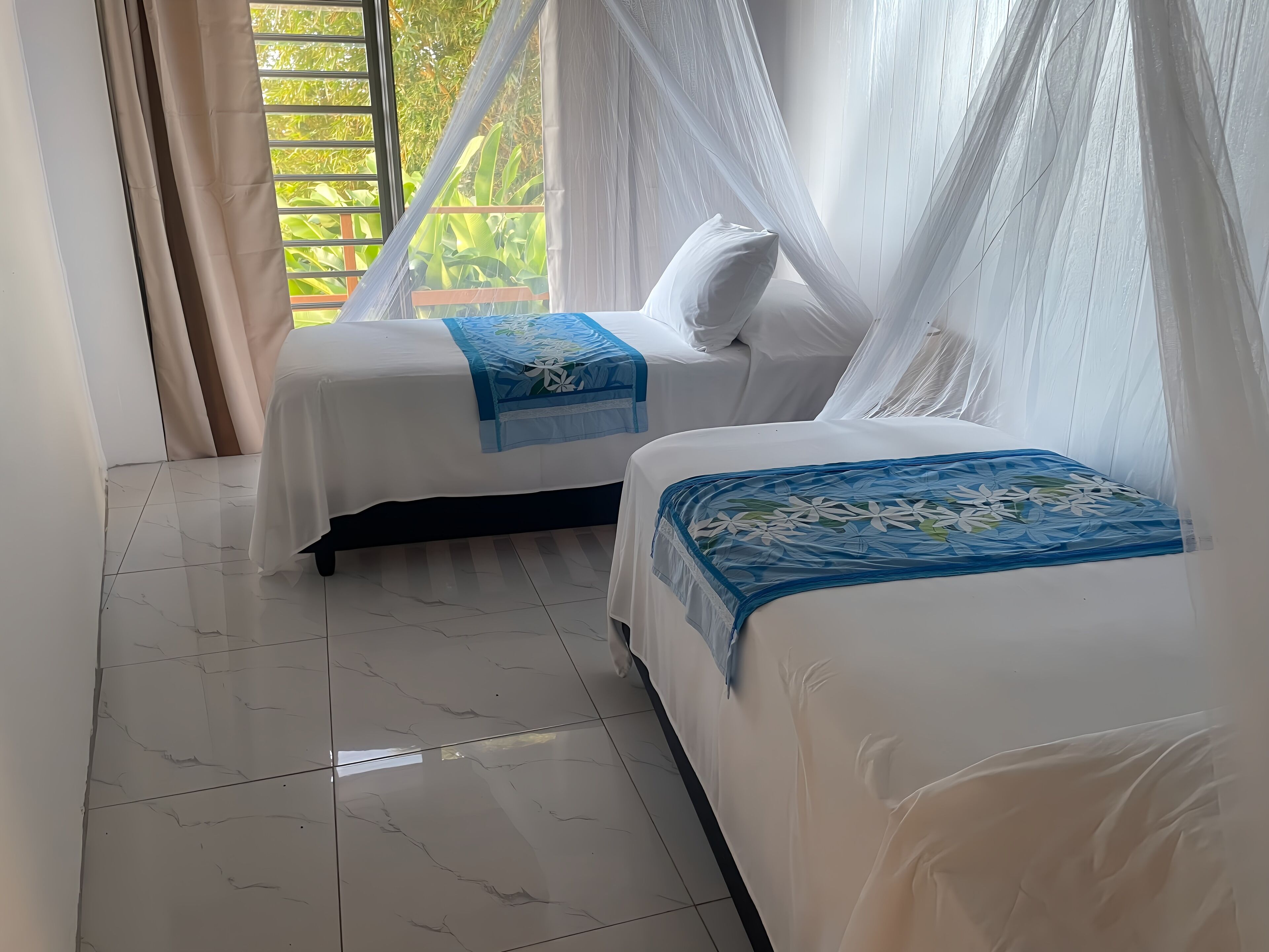 Une chambre avec deux lits, une fenêtre donnant sur un paysage verdoyant et un sol carrelé.